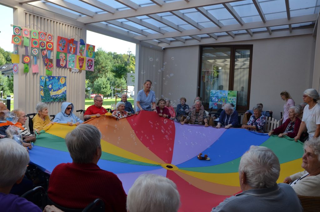 Seniorzy bawią się z chustą animacyjną na tarasie domu seniora.